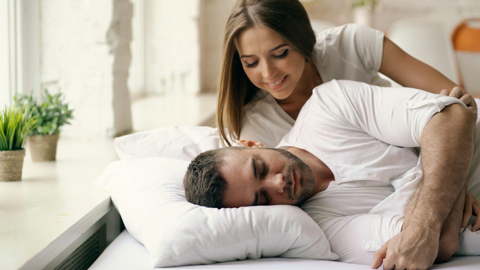 A woman leaning over a man asleep on his side in bed