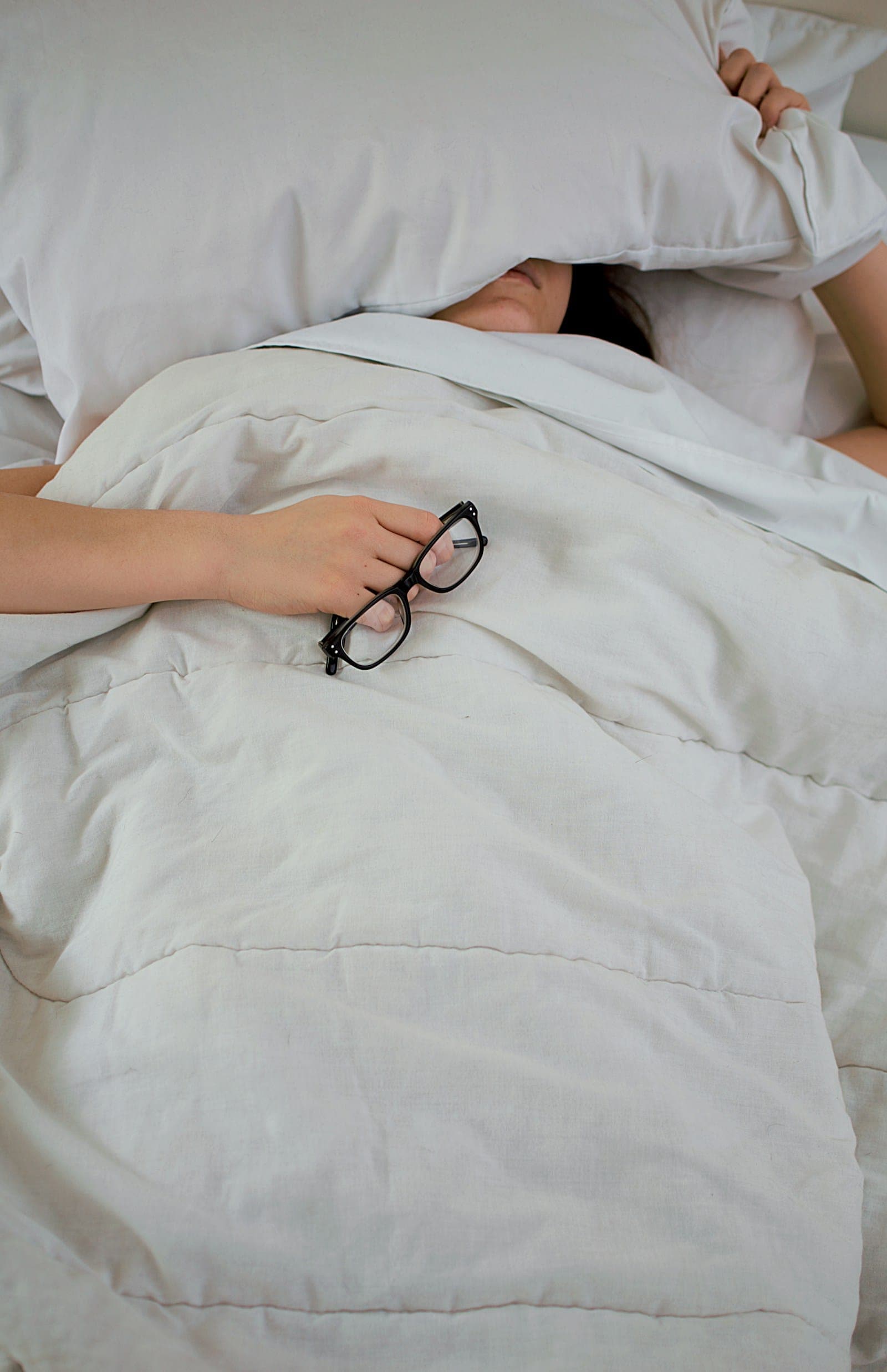 A person lying in bed under a white duvet, holding a pair of glasses